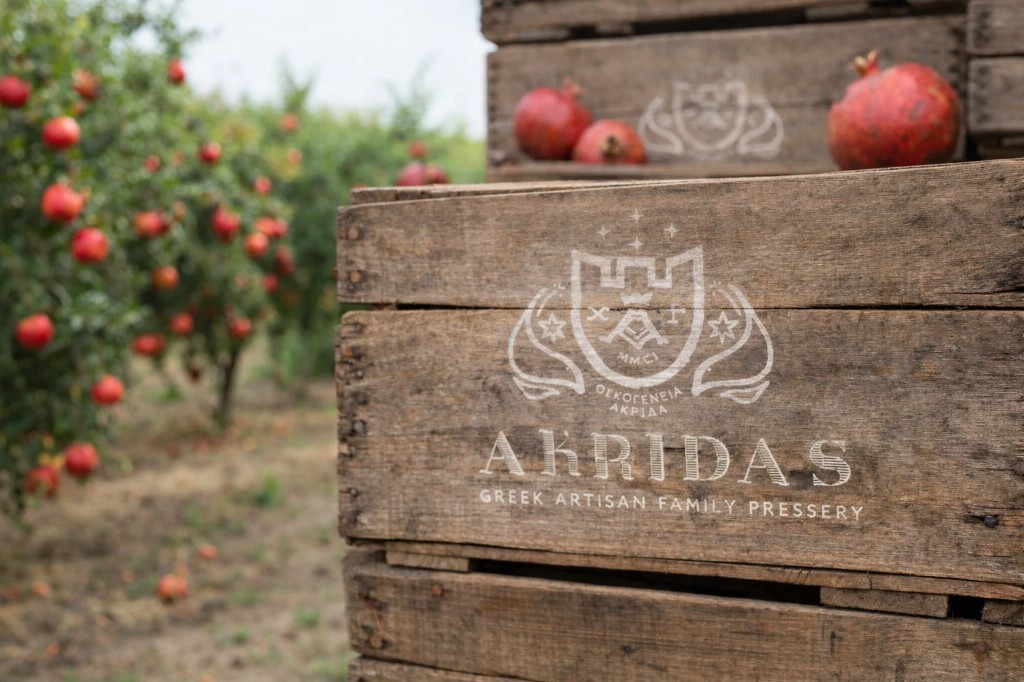 Stacked wooden crates with the Akridas Greek Artisan Family Pressery logo in a pomegranate orchard
